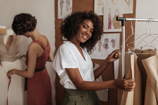 moda sustentável: mulher negra em blusa branca sorri, olha para a câmera e trabalha como estilista com a amiga.